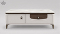 A modern low coffee table with a white faux leather top, featuring a two-drawer front with dark wood inserts and brass-tone hardware, set against a neutral gray background.
